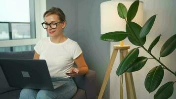 mujer con lentes se sienta en un sofá en un acogedor habitación y hace un en línea compra utilizando un crédito tarjeta y teléfono inteligente en línea compras video