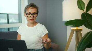 Woman with glasses sits on a couch in a cozy room and makes an online purchase using a credit card and smartphone. Online shopping video