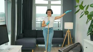 femme dans écouteurs nettoie le maison et avoir amusement en chantant avec une balai comme une étoile à une concert video