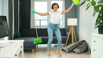 mujer en auriculares limpieza el casa y teniendo divertido bailando con un Escoba y toallita video