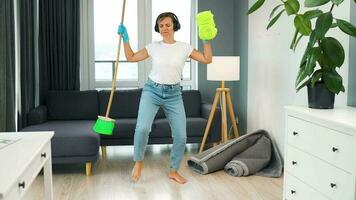 mulher dentro fones de ouvido limpeza a casa e tendo Diversão dançando com uma vassoura e toalha. video