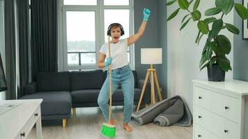 kvinna i hörlurar rengör de hus och ha roligt sång med en kvast tycka om en stjärna på en konsert video
