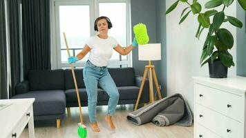 mulher dentro fones de ouvido limpeza a casa e tendo Diversão dançando com uma vassoura e toalha. video