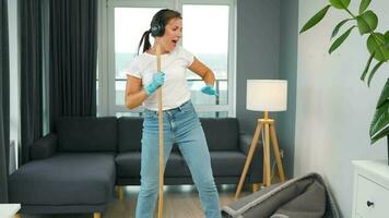 mujer en auriculares limpia el casa y tener divertido canto con un Escoba me gusta un estrella a un concierto video