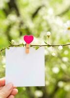 Empty white sheet is attached with a decorative clothespin to a branch against the background of a flowering tree. Mockup. Spring time. Copy space. Selective focus. photo