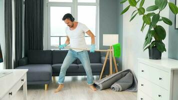 hombre en auriculares limpieza el casa, engañando alrededor y teniendo divertido bailando y canto con un Escoba video