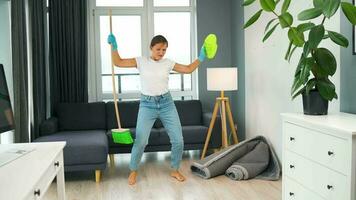 mujer en auriculares limpieza el casa y teniendo divertido bailando con un Escoba y toallita video