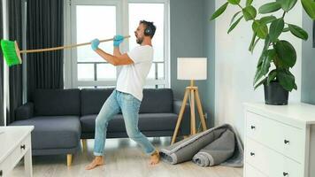 hombre en auriculares limpieza el casa, engañando alrededor y teniendo divertido bailando y canto con un Escoba video