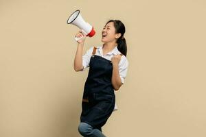 Start a small business. Young Asian businesswoman in white shirt and apron with shouting into megaphone to announce promotion isolated on light brown background. photo