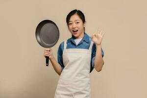 Shocked Asian woman in apron standing isolated on light brown background. She made an OK gesture and was holding a frying pan in her hand. photo