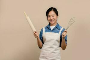 Cheerful Asian woman wearing apron and holding Baking equipment in hand standing isolated on light brown background. photo