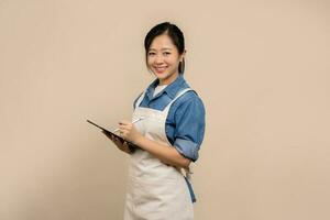 Cheerful asian woman in apron with a tablet in hand isolated on background in studio. Small business startup concept. photo