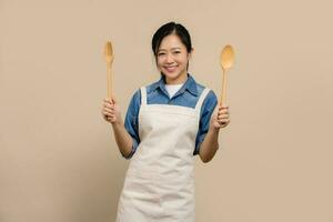 Portrait cheerful asian woman wearing apron and holding fork spoon on light brown background. photo