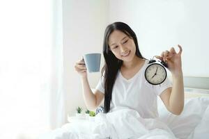 hermosa asiático mujer con largo negro pelo y en en blanco pijama. ella participación alarma reloj y un café taza en el cama y estaba despertar arriba en el Mañana. ella sintió muy renovado foto