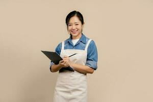 Charming young Asian businesswoman owner wearing an apron holding and holding clipboard front of an isolated over light brown background. photo