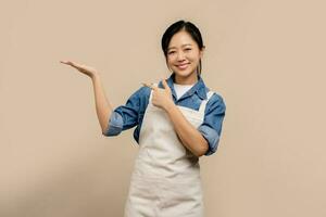 Small business owner of young asian woman in apron and pointing hand aside isolated on a light brown background. Small business startup concept photo