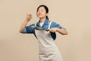 Young Asian housewife in apron and holding pan standing isolated on light brown background. She was inhaling the aroma of food in the pan. photo