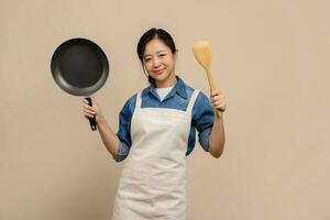 Young Asian woman housewife wearing kitchen apron cooking and holding pan and spatula isolated on light brown background. photo