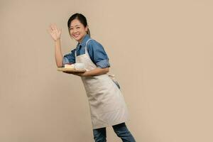 Asian barista waitress in casual clothes and apron She is holding a cake and a hot drink on a wooden tray on a light brown background. photo