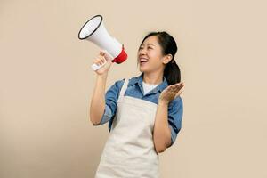 Beautiful Asian woman in apron with megaphone in hand. She is shouting excitedly standing isolated on a light brown background. photo