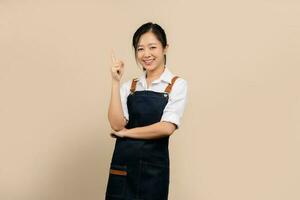 Happy young asian businesswoman wearing white shirt and apron with copy space on light brown background. photo