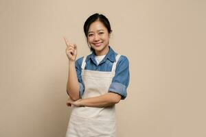 Smiling asian young woman wearing apron uniform hand gesture finger point up isolated on background photo
