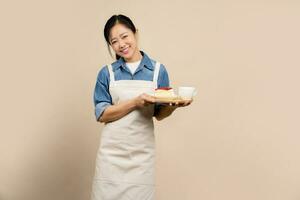Young Asian waitress wearing apron on light brown background. She is happily holding a cake and a hot drink on the top wooden tray. photo