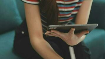 contento asiatico adolescente ragazza Tenere tampone computer aggeggio utilizzando digitale tavoletta tecnologia seduta su il divano a casa. sorridente giovane donna utilizzando app, shopping in linea, lettura notizia, navigazione Internet su divano video