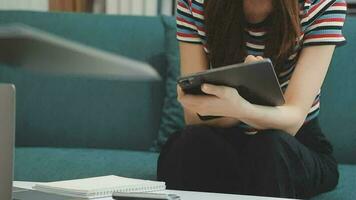 Happy Asian teen girl holding pad computer gadget using digital tablet technology sitting on the couch at home. Smiling young woman using apps, shopping online, reading news, browsing internet on sofa video