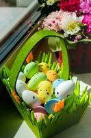 Easter green basket with plastic colored eggs on the windowsill against the background of flowers photo