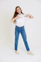 linda y sonriente niña en un blanco camiseta señalando abajo o a su camiseta, sonriente alegremente y expresando positivo emociones en un blanco antecedentes. foto