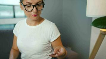 vrouw met bril zit Aan een bankstel in een knus kamer en maakt een online aankoop gebruik makend van een credit kaart en smartphone. online boodschappen doen video