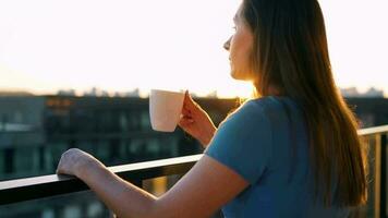 mulher com uma copo do café em pé em a sacada e admirar a pôr do sol video