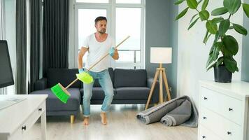 hombre en auriculares limpieza el casa, engañando alrededor y teniendo divertido bailando y canto con un Escoba video