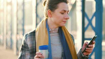 portrait de une Jeune caucasien femme d'affaires dans une manteau, en marchant à travers le pont sur une glacial ensoleillé matin, en buvant café et en utilisant téléphone intelligent. communication, travail jour, occupé la vie concept video