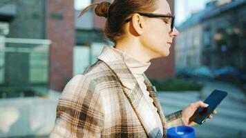 retrato de un joven caucásico mujer de negocios con lentes y un Saco camina mediante el negocio distrito, Bebiendo café y utilizando teléfono inteligente video