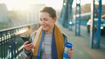 porträtt av en ung caucasian affärskvinna i en täcka, gående tvärs över de bro på en frostig solig morgon, dricka kaffe och använder sig av smartphone. kommunikation, arbete dag, upptagen liv begrepp video