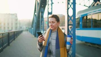 portret van een jong Kaukasisch zakenvrouw in een jas, wandelen aan de overkant de brug Aan een ijzig zonnig ochtend, drinken koffie en gebruik makend van smartphone. communicatie, werk dag, bezig leven concept video