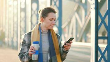 portret van een jong Kaukasisch zakenvrouw in een jas, wandelen aan de overkant de brug Aan een ijzig zonnig ochtend, drinken koffie en gebruik makend van smartphone. communicatie, werk dag, bezig leven concept video