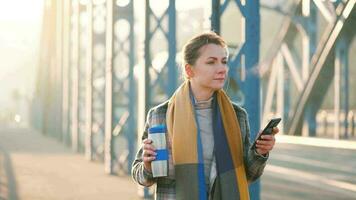 portret van een jong Kaukasisch zakenvrouw in een jas, wandelen aan de overkant de brug Aan een ijzig zonnig ochtend, drinken koffie en gebruik makend van smartphone. communicatie, werk dag, bezig leven concept video