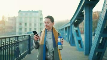 portret van een jong Kaukasisch zakenvrouw in een jas, wandelen aan de overkant de brug Aan een ijzig zonnig ochtend, drinken koffie en gebruik makend van smartphone. communicatie, werk dag, bezig leven concept video