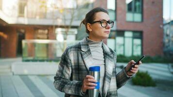 retrato de un joven caucásico mujer de negocios con lentes y un Saco camina mediante el negocio distrito, Bebiendo café y utilizando teléfono inteligente video