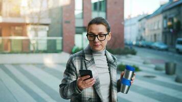 retrato de un joven caucásico mujer de negocios con lentes y un Saco camina mediante el negocio distrito, Bebiendo café y utilizando teléfono inteligente video