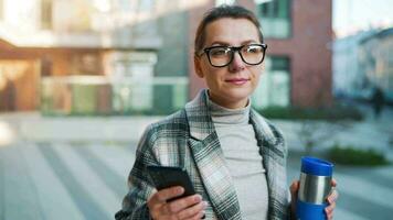 portret van een jong Kaukasisch zakenvrouw met bril en een jas wandelingen door de bedrijf wijk, drinken koffie en gebruik makend van smartphone video