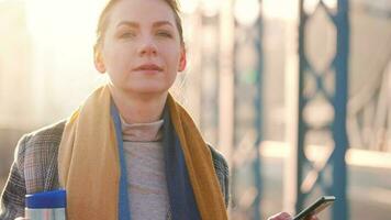 portrait de une Jeune caucasien femme d'affaires dans une manteau, en marchant à travers le pont sur une glacial ensoleillé matin, en buvant café et en utilisant téléphone intelligent. communication, travail jour, occupé la vie concept video