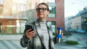 Porträt von ein jung kaukasisch Geschäftsfrau mit Brille und ein Mantel Spaziergänge durch das Geschäft Bezirk, Trinken Kaffee und mit Smartphone video