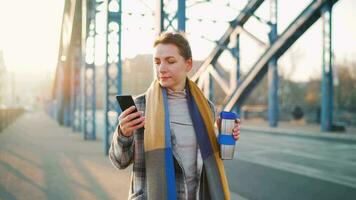 portret van een jong Kaukasisch zakenvrouw in een jas, wandelen aan de overkant de brug Aan een ijzig zonnig ochtend, drinken koffie en gebruik makend van smartphone. communicatie, werk dag, bezig leven concept video