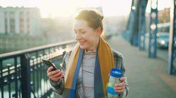retrato do uma jovem caucasiano empresária dentro uma casaco, caminhando através a ponte em uma gelado ensolarado manhã, bebendo café e usando Smartphone. comunicação, trabalhos dia, ocupado vida conceito video