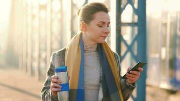 portret van een jong Kaukasisch zakenvrouw in een jas, wandelen aan de overkant de brug Aan een ijzig zonnig ochtend, drinken koffie en gebruik makend van smartphone. communicatie, werk dag, bezig leven concept video