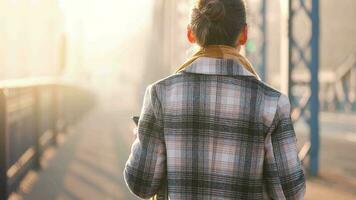 retrato de un joven caucásico mujer de negocios en un abrigo, caminando a través de el puente en un escarchado soleado mañana, Bebiendo café y utilizando teléfono inteligente comunicación, trabajo día, ocupado vida concepto video